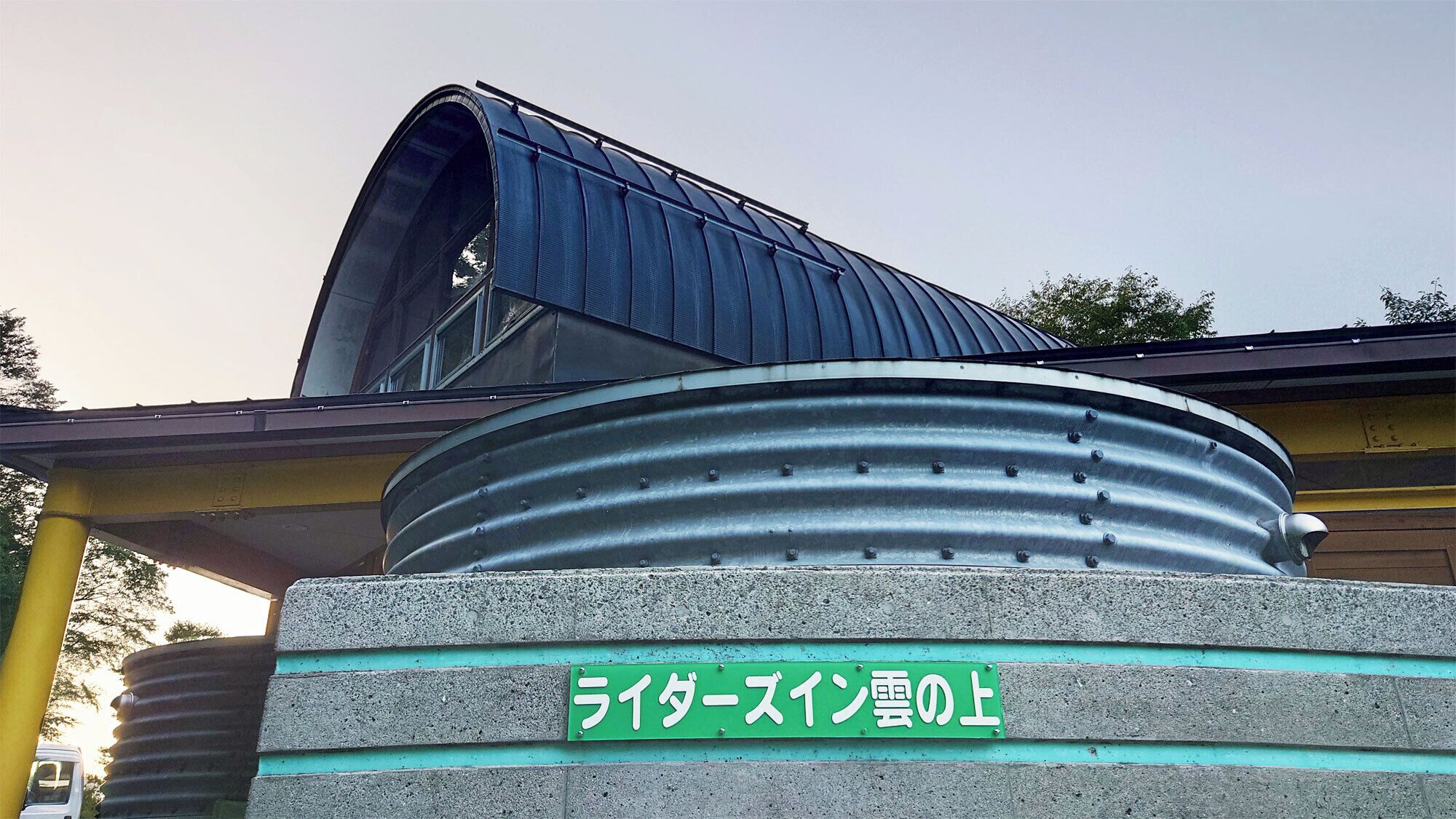 A building with a curved metal structure, a green sign with Japanese characters, and a concrete base.