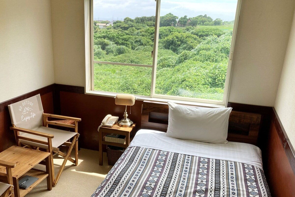 Una habitación con cama, escritorio, silla, mesita de noche con lámpara y ventana con vista a zonas verdes.
