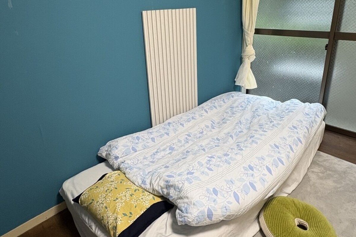 Una cama individual con colcha floral, almohada blanca, almohada amarilla, cojín verde, cabecera blanca, un reloj en la pared y una ventana con cortina.