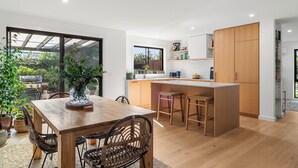 A modern kitchen with wooden cabinets, a central island, and a dining area with a wooden table and chairs. - Central and very close to retail hub, restaurants, cafes, Coles, Woolworths (Cowes)