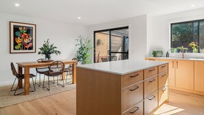 A modern kitchen with wooden cabinets, a white countertop, and a dining area with a table and chairs. - Central and very close to retail hub, restaurants, cafes, Coles, Woolworths (Cowes)