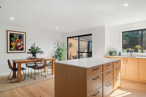 A modern kitchen with wooden cabinets, a white countertop, and a dining area with a table and chairs. - Central and very close to retail hub, restaurants, cafes, Coles, Woolworths (Cowes)