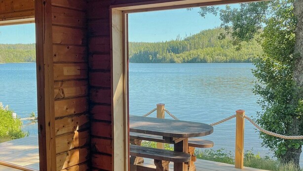 Una cabaña con interior de madera, un ventanal grande con vista a un lago, una mesa de picnic afuera y una chimenea en el interior.