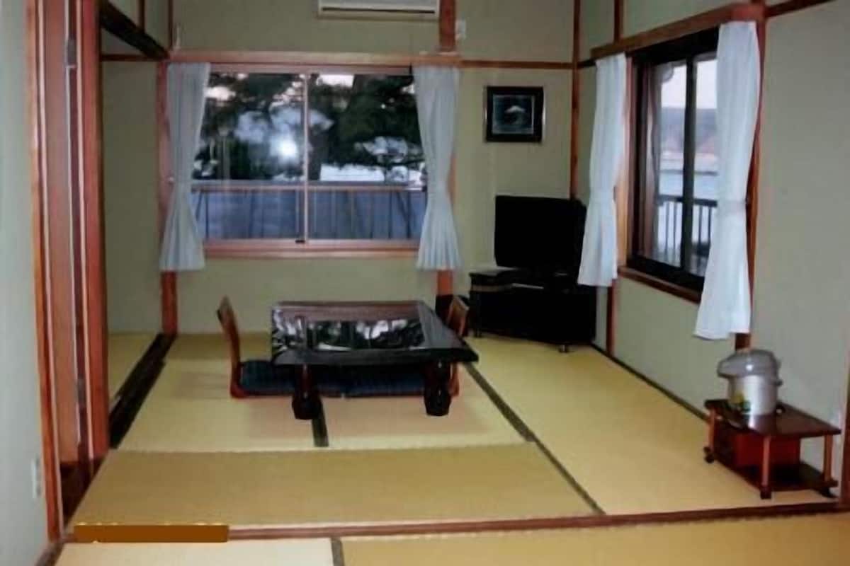 Ein traditioneller japanischer Raum mit Tatami-Böden, einem niedrigen Tisch, einem Fernseher und einem Fenster mit Vorhängen.