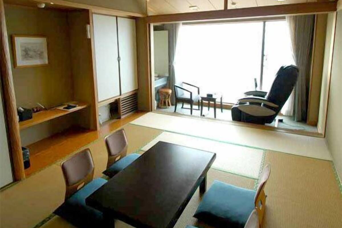 Una habitación con suelo de tatami, una mesa con sillas, un sofá, una ventana con cortinas y una pequeña zona de estar con una silla y una mesa.