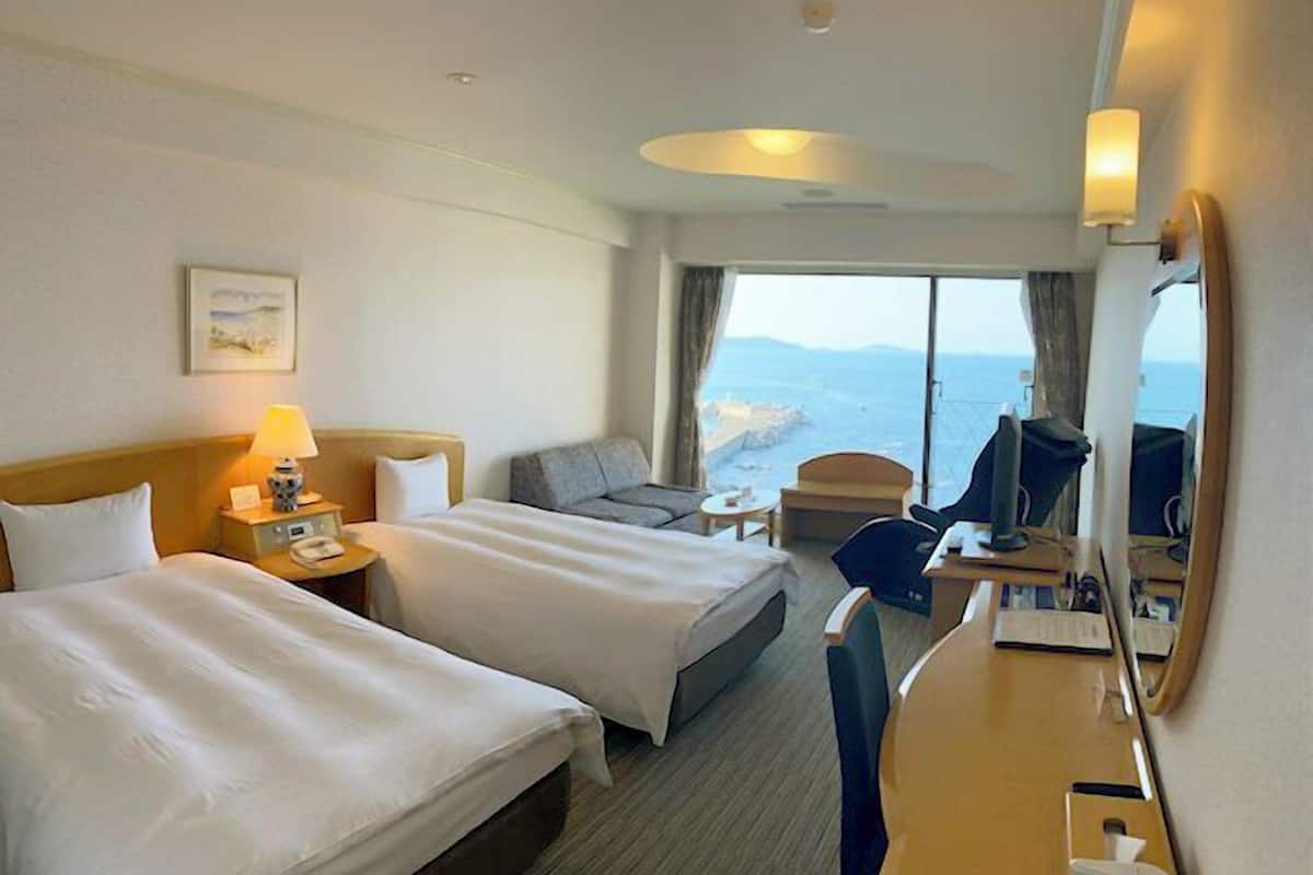A hotel room with two beds, a desk, a chair, a lamp, a phone, a mirror, and a view of the sea.