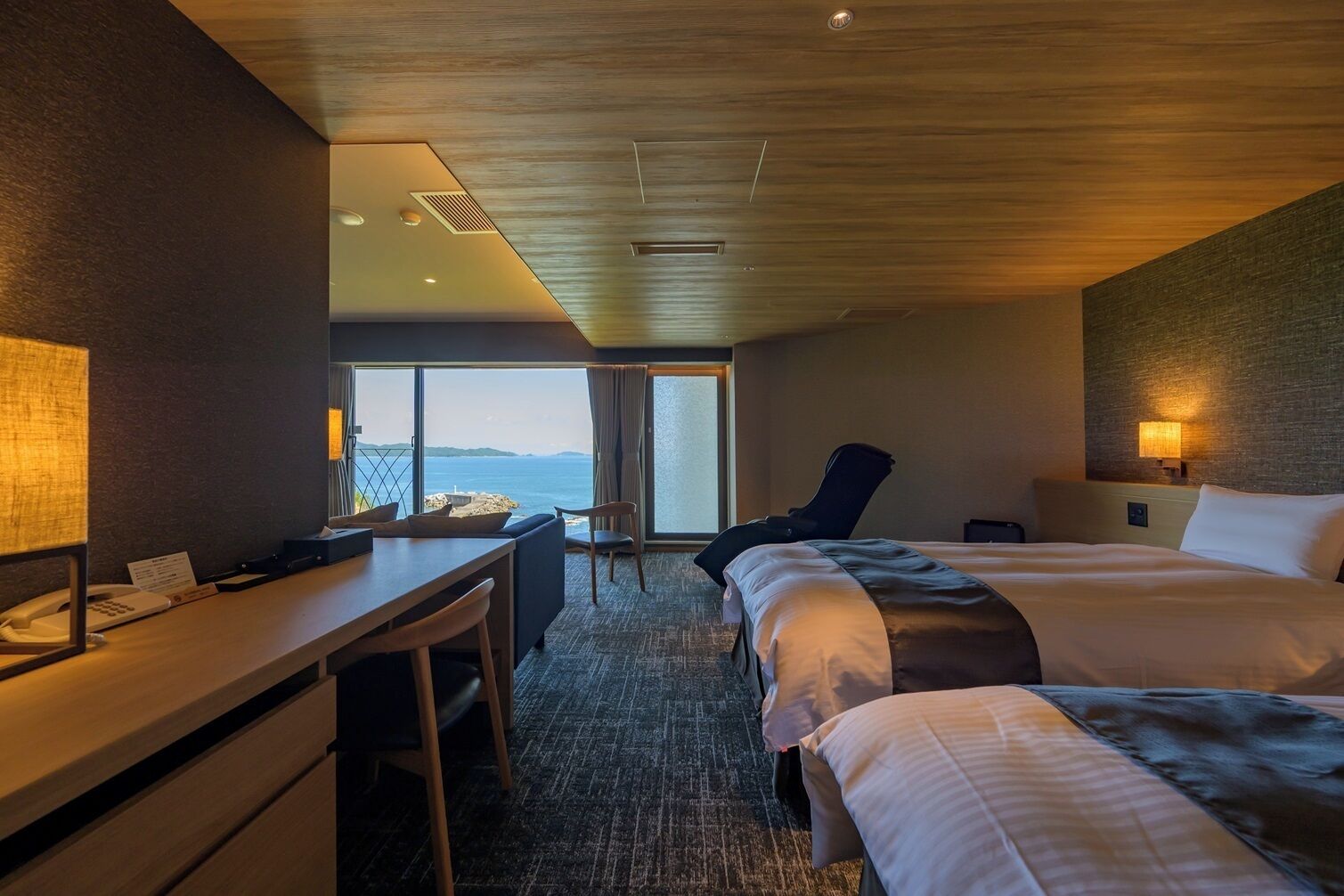 A hotel room with a desk, two beds, a chair, a view of the sea, and a wooden ceiling.