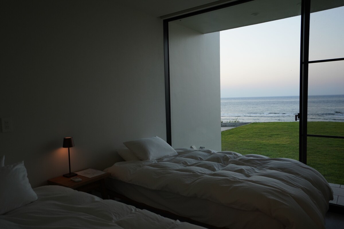A bedroom with a bed, a nightstand, a lamp, and a large window offering a view of the ocean.