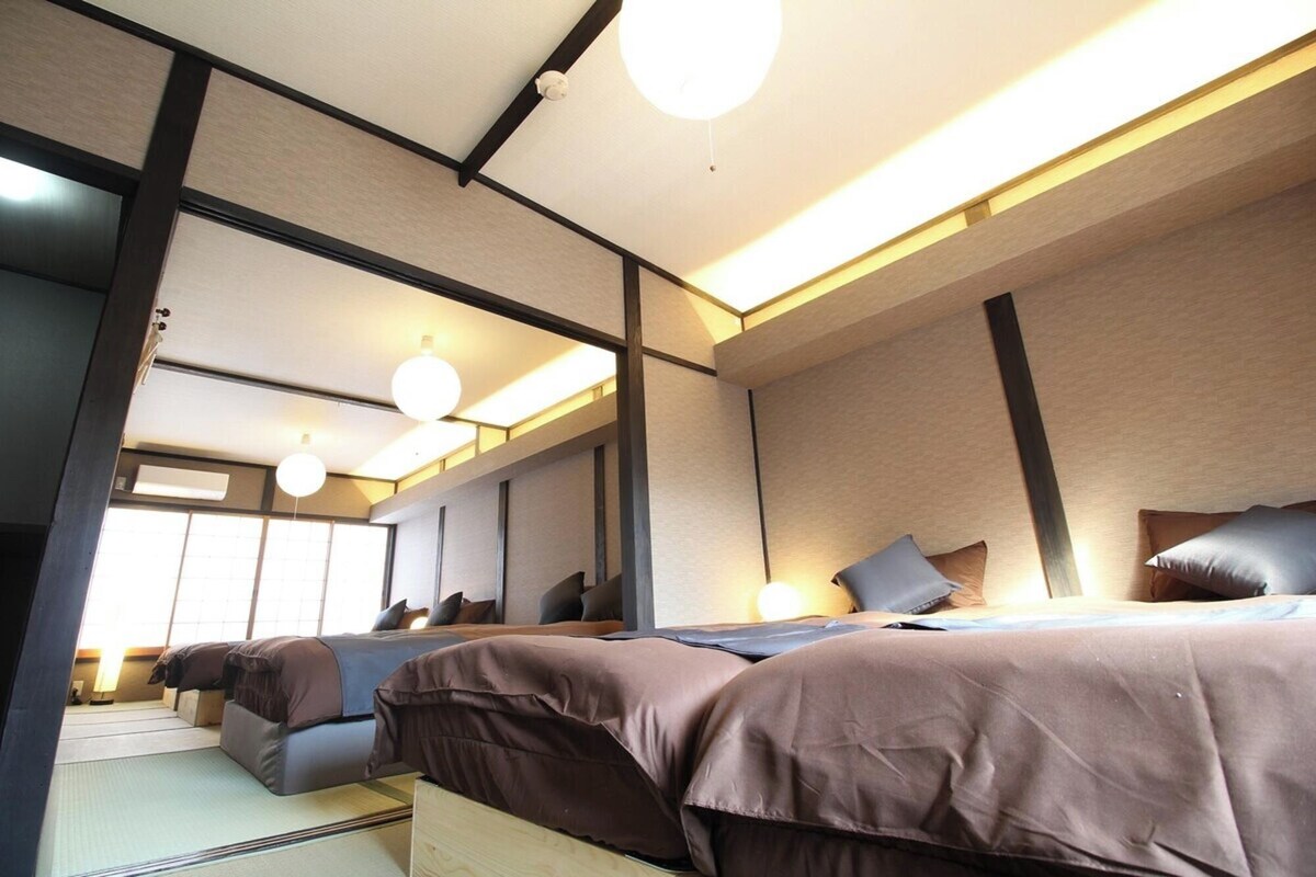 Ett modernt hotellrum med en stor säng, en mindre säng och en soffa. Det finns flera kuddar och ett sängbord. Rummet har ett stort fönster och en taklampa.