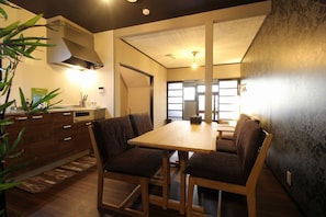 A dining area with a wooden table and chairs, a kitchen in the background, and a large window allowing natural light. - Kyo-hiiragi (Kyoto)