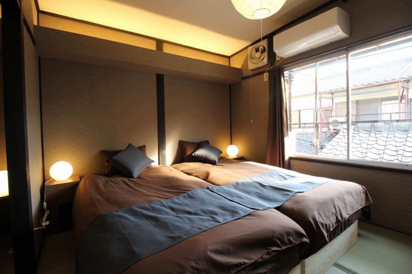 A bedroom with a bed, bedside lamps, an air conditioning unit, and a window with a view of buildings.