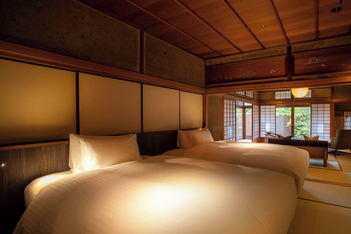 A traditional Japanese-style bedroom with tatami flooring, a large bed with white bedding, sliding shoji doors, and a view of greenery outside.