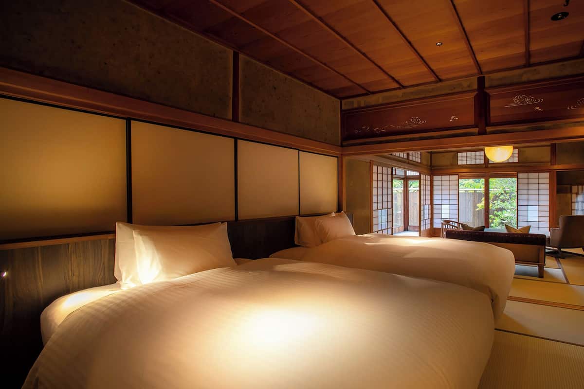 A traditional Japanese-style bedroom with tatami flooring, a large bed with white bedding, sliding shoji doors, and a view of greenery outside.