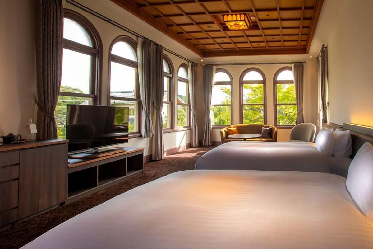 A hotel room with a large bed, a TV, a desk, and a window with a view of greenery.