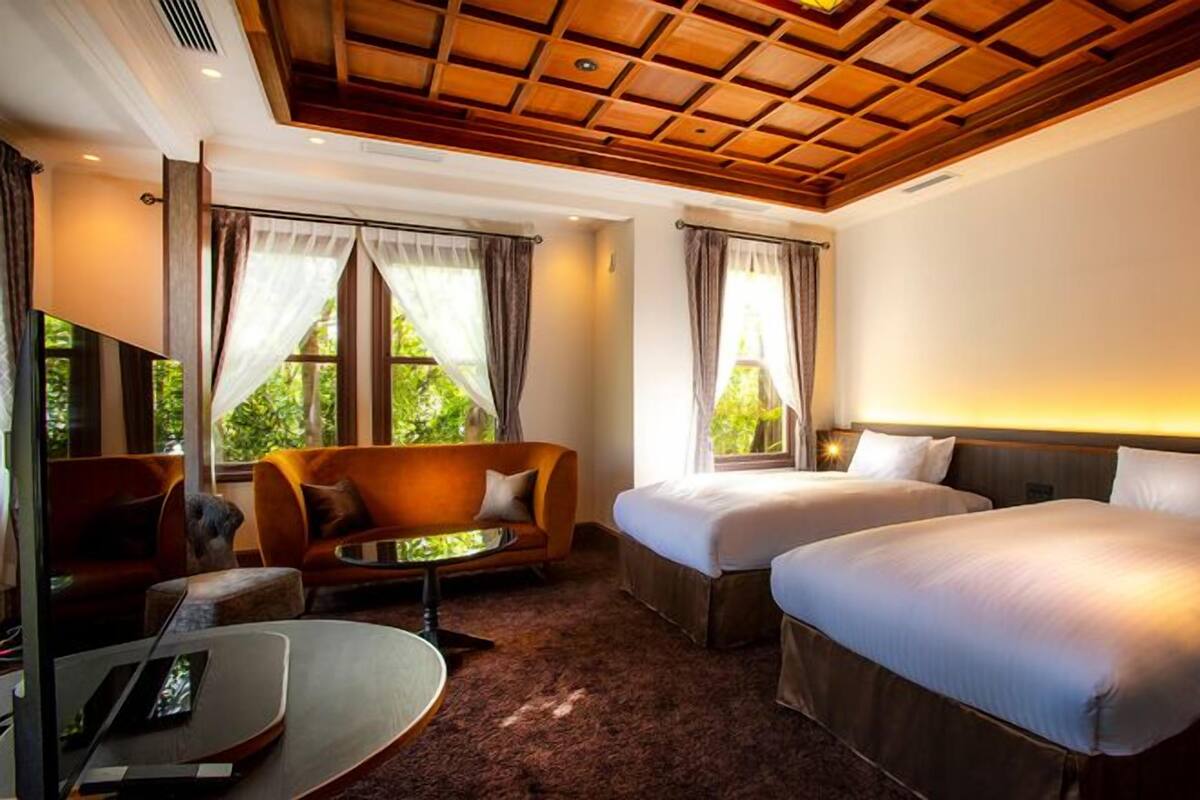A hotel room with two beds, a sofa, a TV, and large windows.