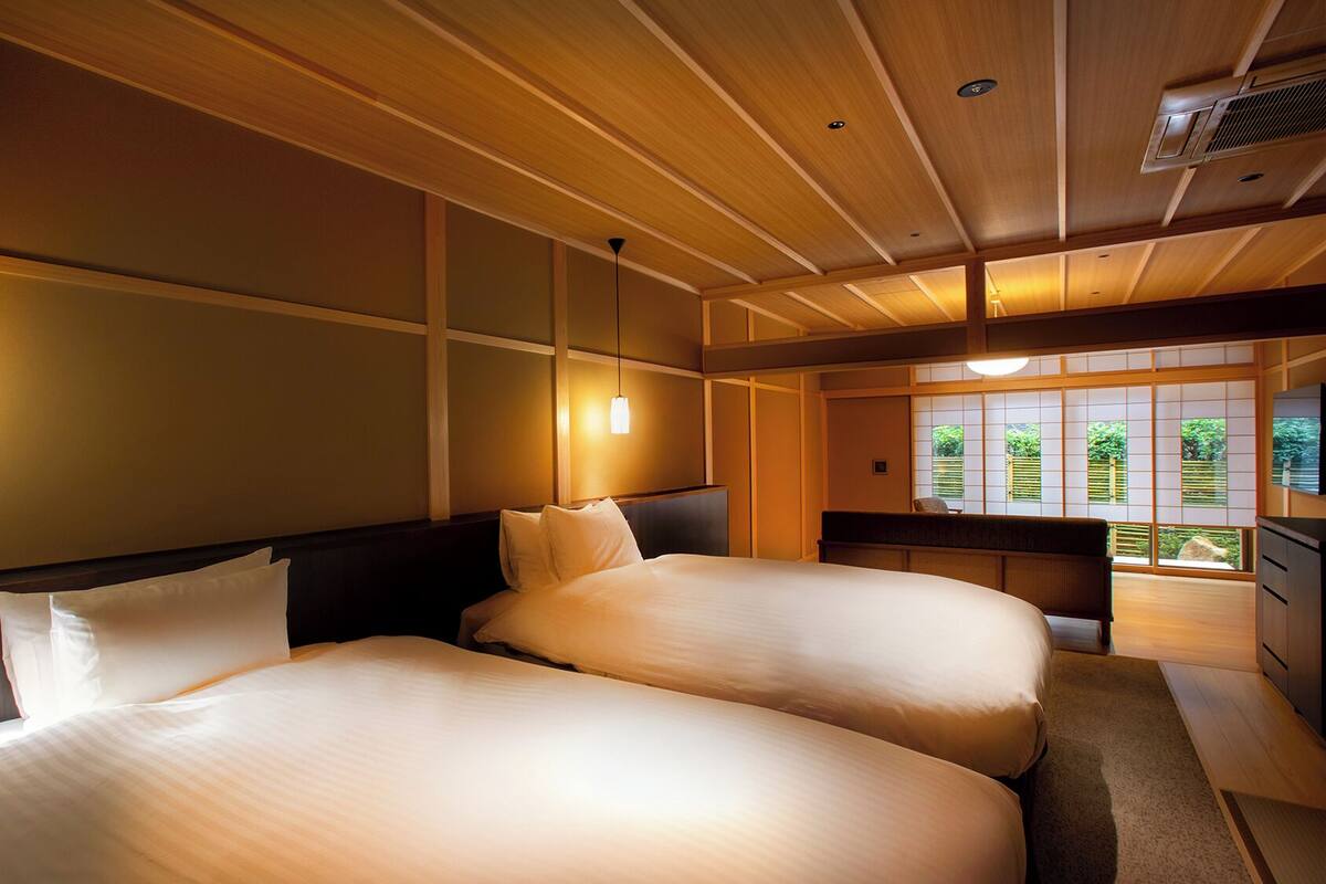 A hotel room with two beds, wooden ceiling, and large windows with a view of greenery.
