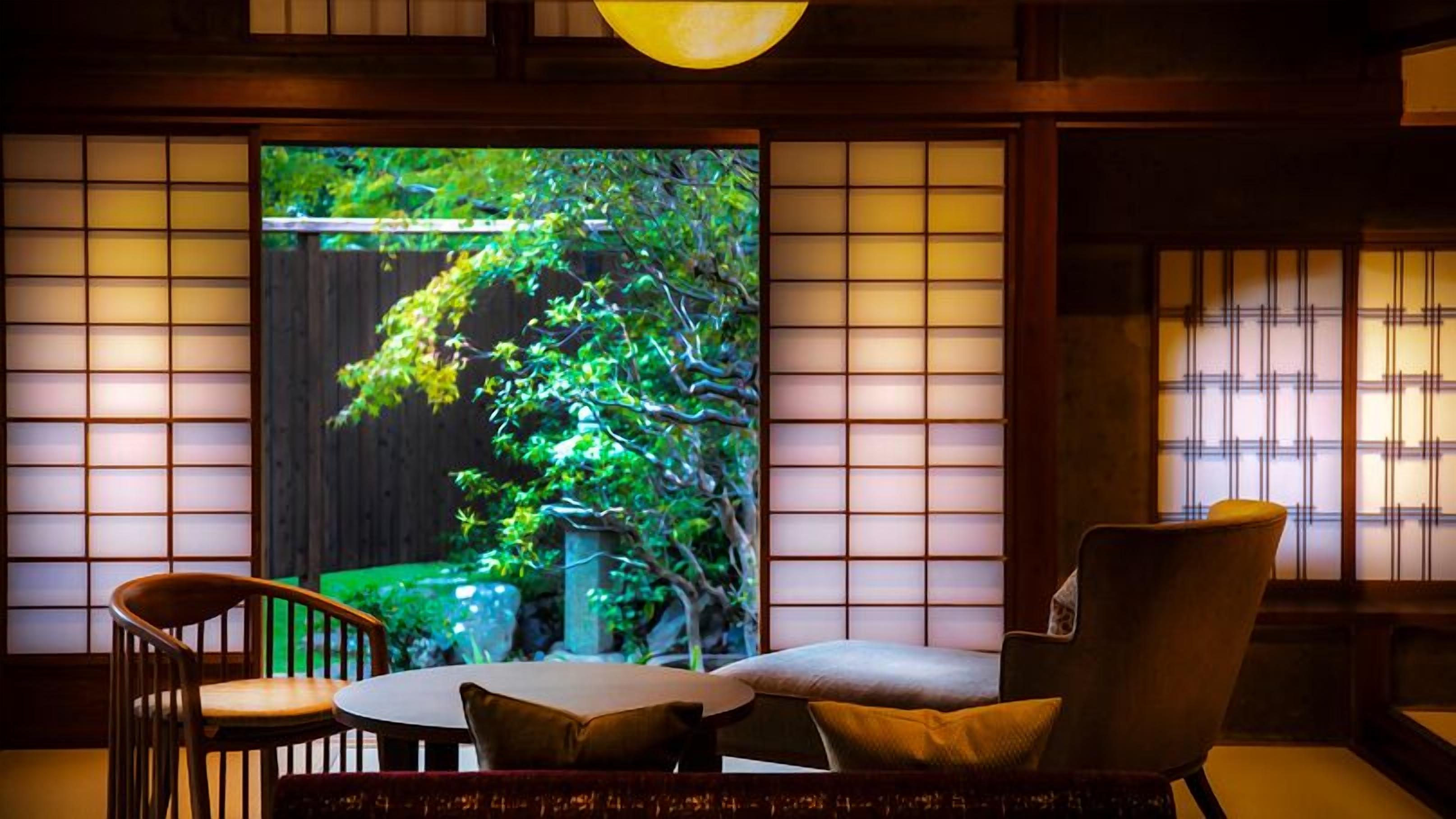 A traditional room with tatami flooring, a low table, a cushioned chair, and a large sliding door with a view of greenery.