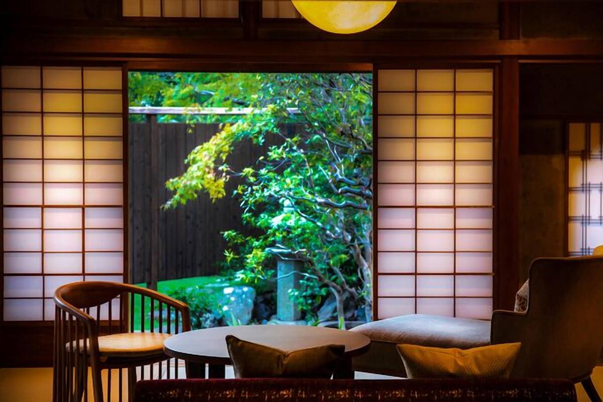 A traditional room with tatami flooring, a low table, a cushioned chair, and a large sliding door with a view of greenery.
