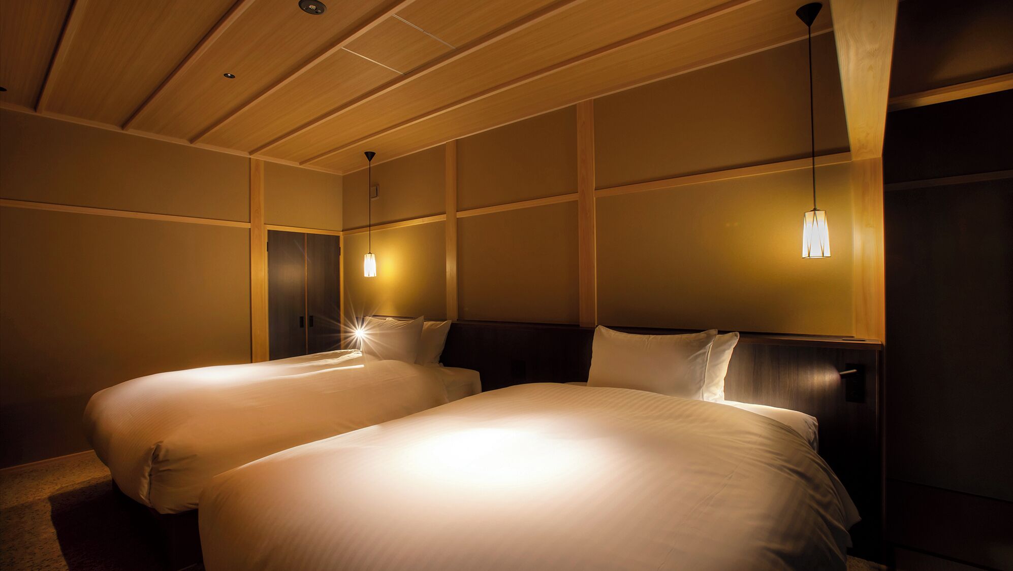 A hotel room with two beds, wooden paneling on the walls, and pendant lights.