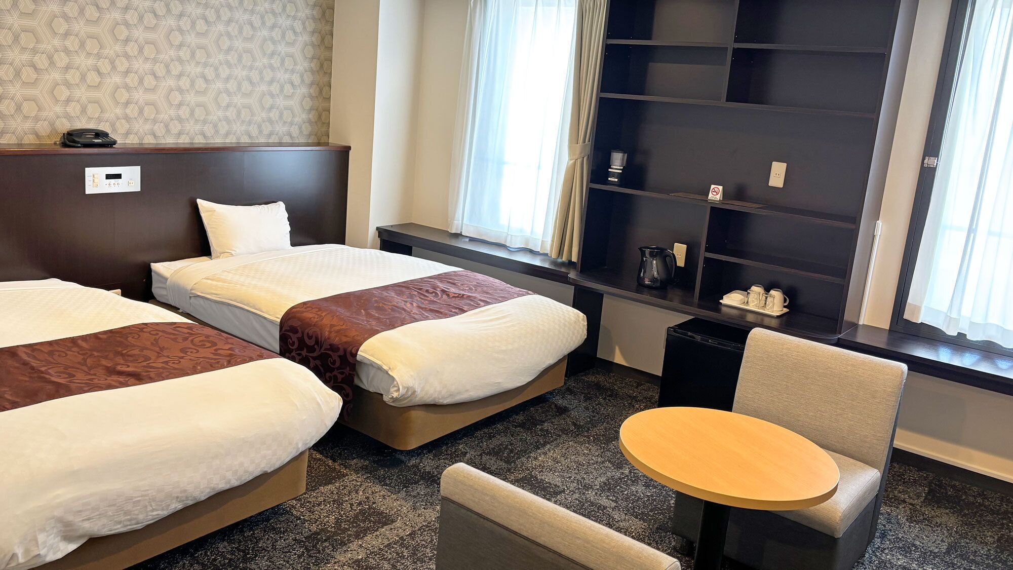 Quarto de hotel com duas camas, uma mesinha, uma cadeira, uma prateleira com amenities de higiene pessoal e uma janela com cortinas.