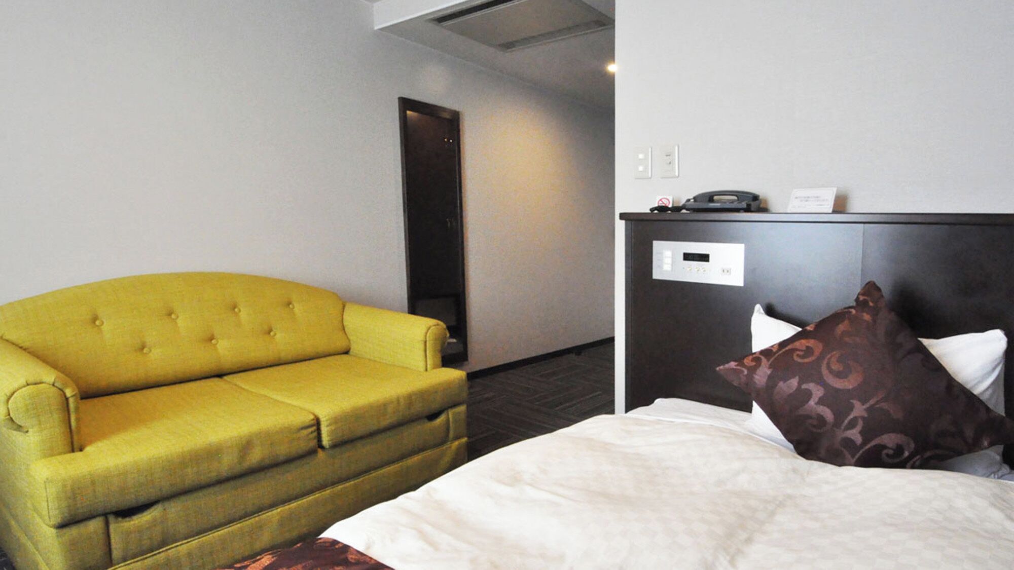A hotel room with a yellow sofa, a bed with a white comforter and brown pillows, a dark headboard, and a small table with a telephone and a sign.