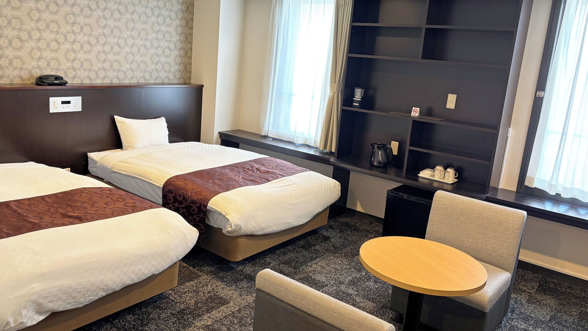 A hotel room with two beds, a small table, a chair, a minibar, and a window with curtains.