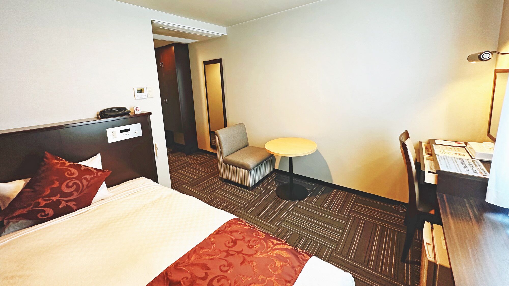 Quarto de hotel com cama, uma cadeira, uma mesinha e uma escrivaninha com telefone.