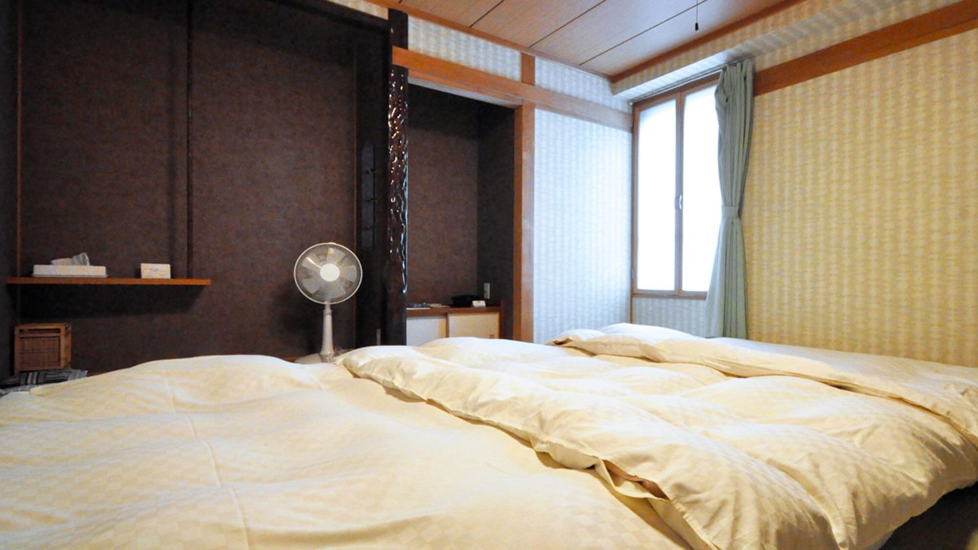 Quarto de hotel com cama, ventilador, janela com cortinas e prateleira com objetos.