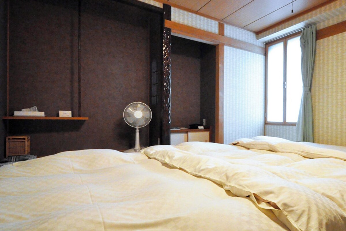 Una habitación de hotel con una cama, un ventilador, una ventana con cortinas y una estantería con objetos.