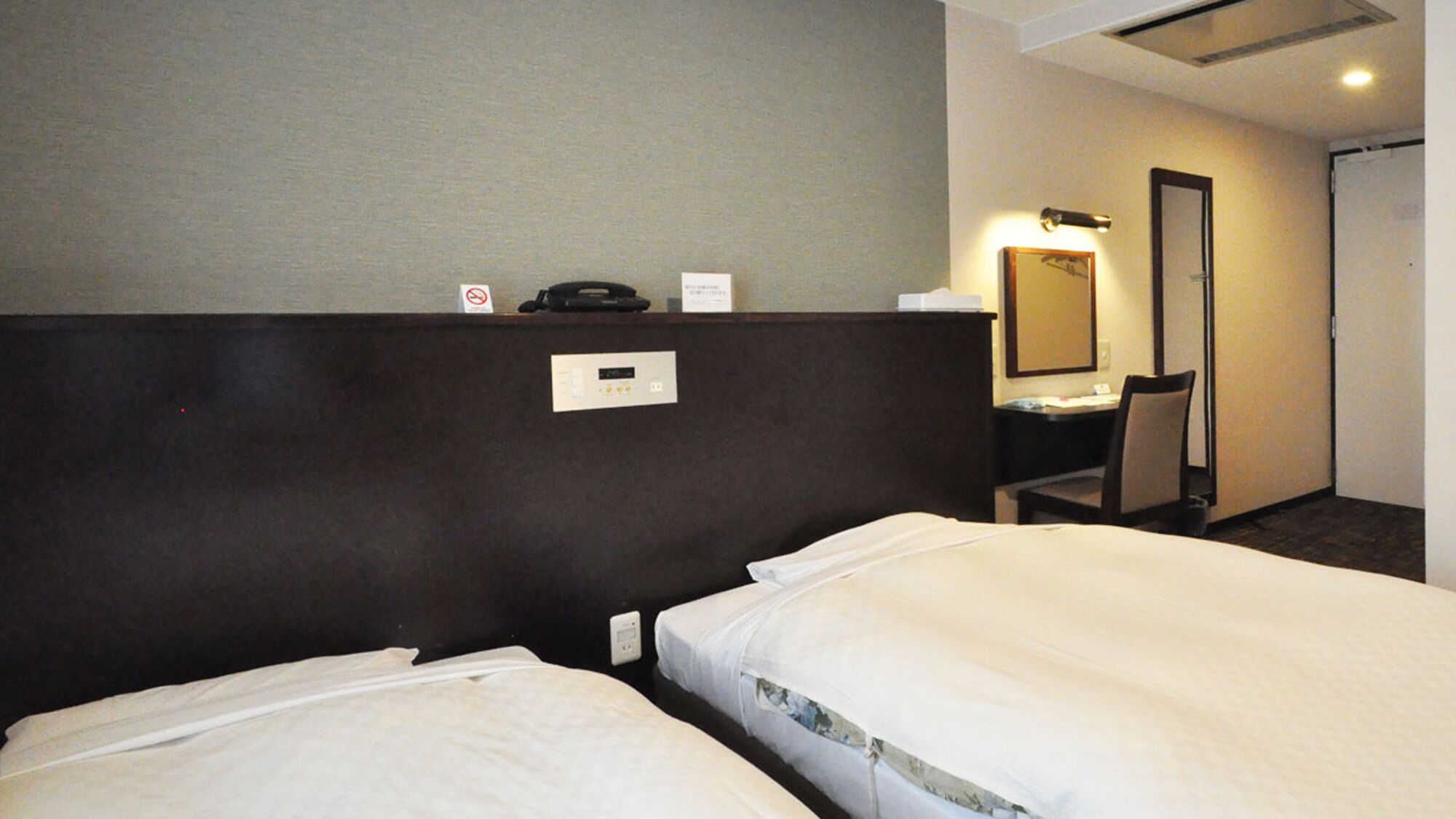 Quarto de hotel com duas camas, uma escrivaninha, uma cadeira, uma luminária de mesa, um espelho e uma porta.