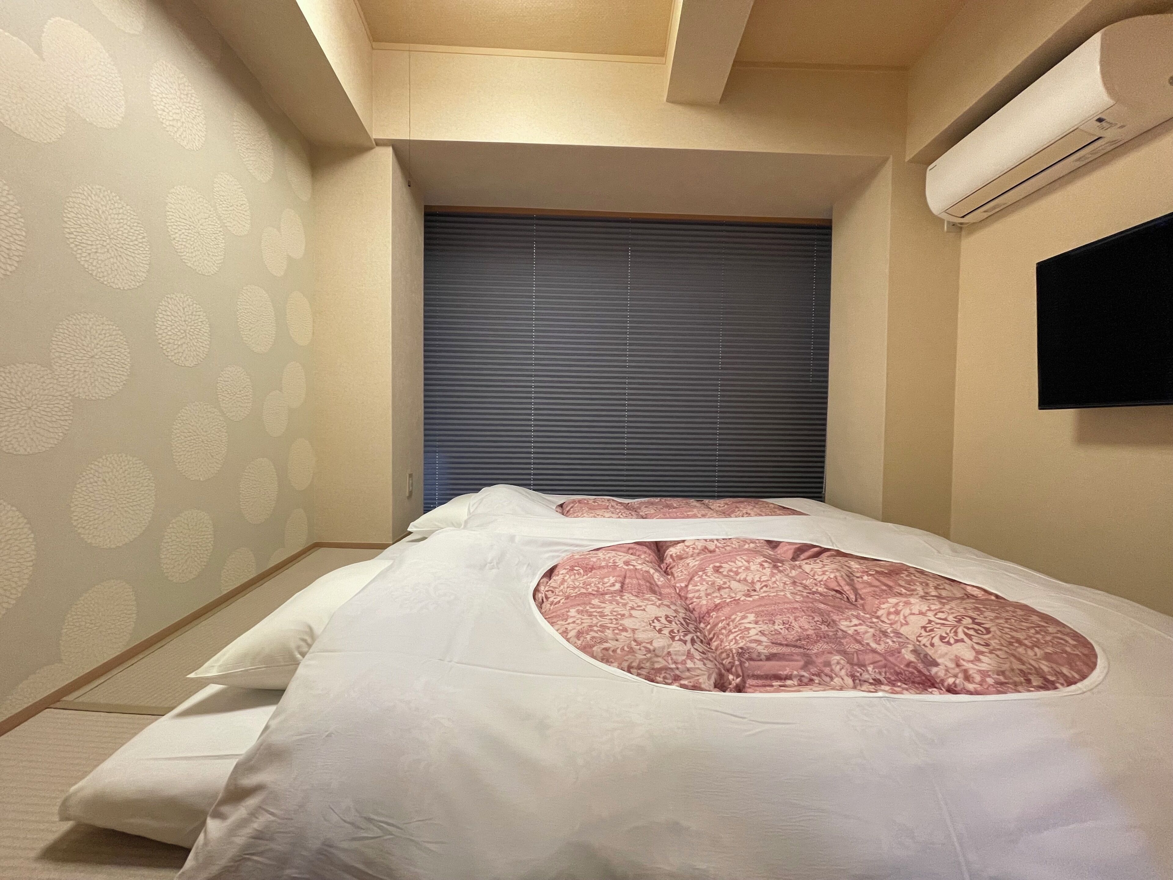 A bedroom with a bed, a window with blinds, an air conditioning unit, and a wall-mounted TV.