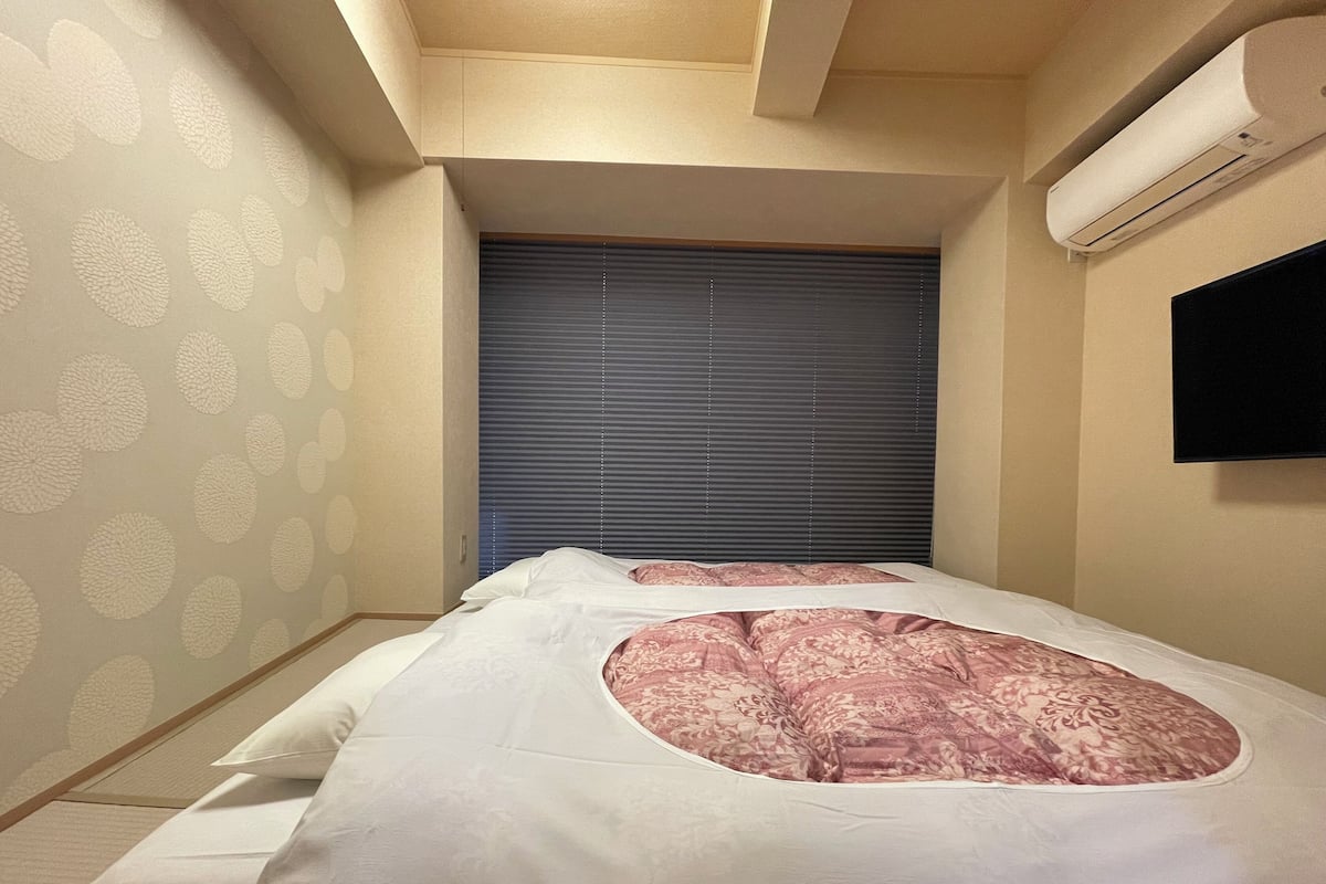 A bedroom with a bed, a window with blinds, an air conditioning unit, and a wall-mounted TV.