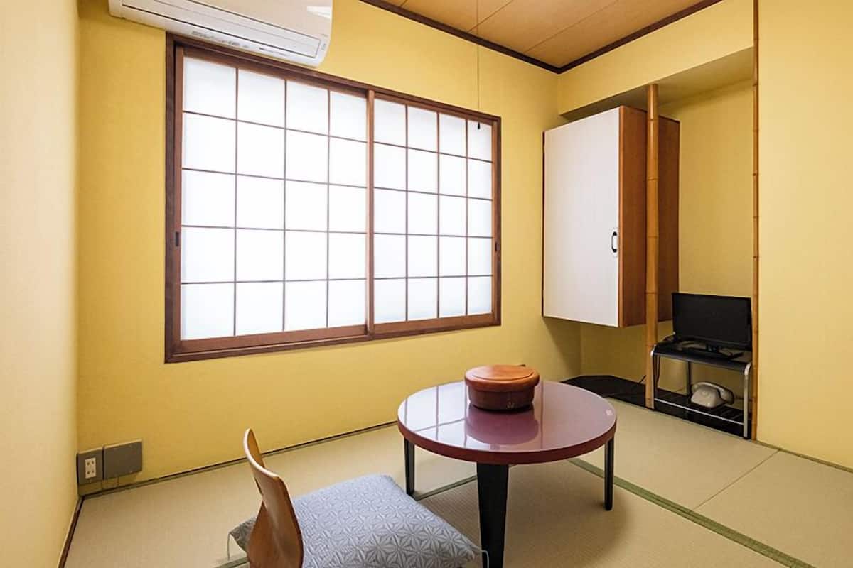 A room with tatami flooring, a low table with a cushion, a sliding door, and a small kitchenette.