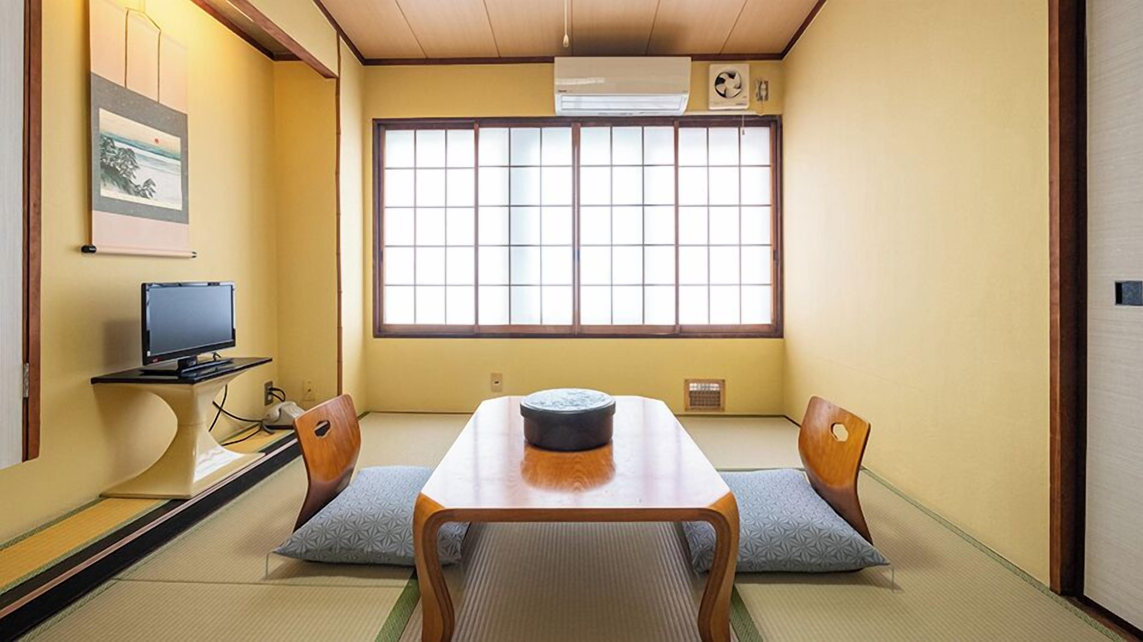 A traditional Japanese room with tatami flooring, a low table, two chairs, a television, and a large window with a view.