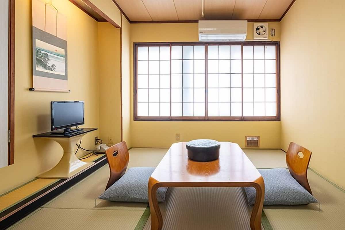 A traditional Japanese room with tatami flooring, a low table with a bowl, two chairs, a television, and a large window.