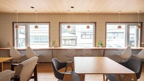 Een moderne eetruimte met houten tafels en stoelen, grote ramen met uitzicht op gebouwen, en hanglampen.