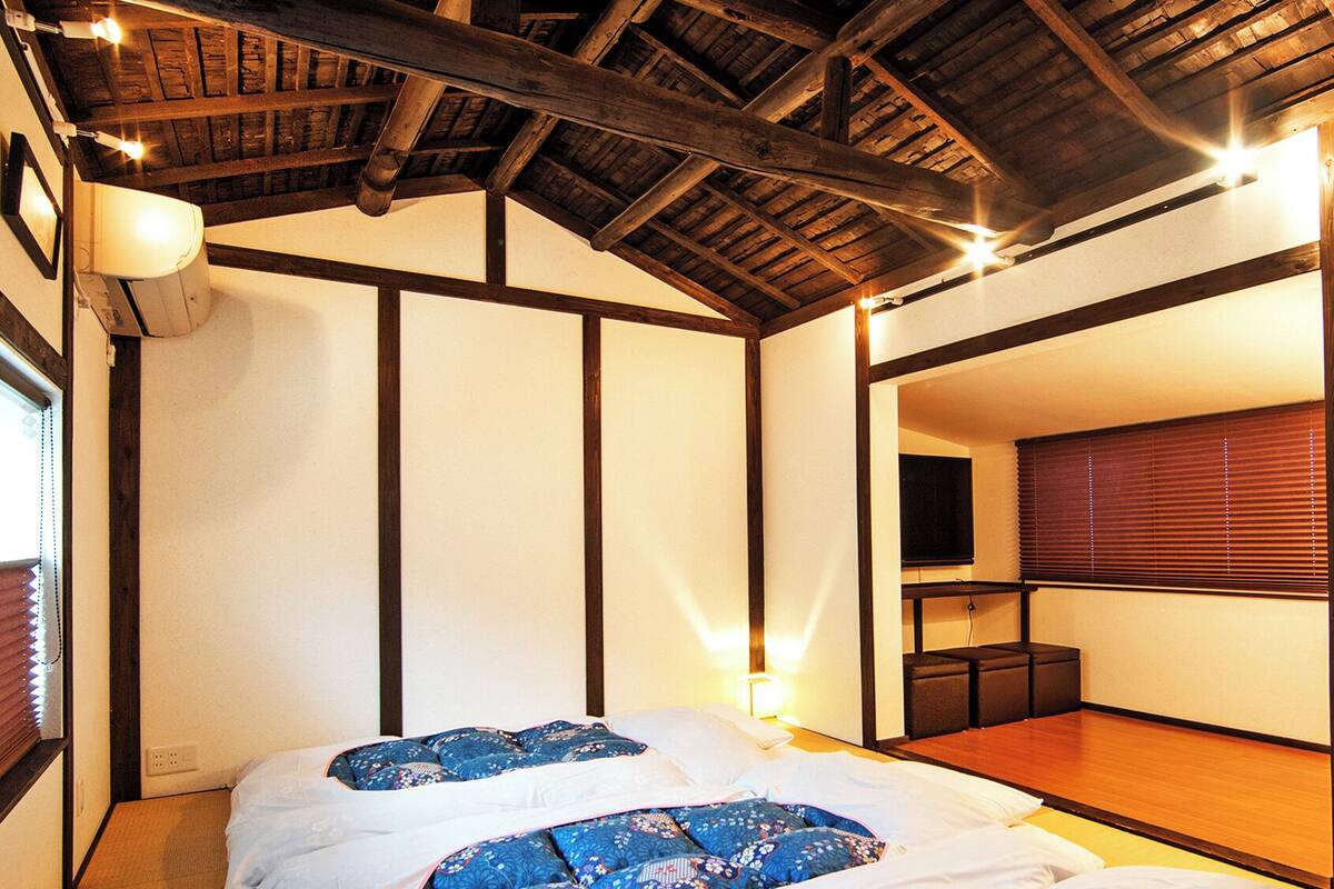 Ein traditioneller japanischer Raum mit Tatami-Boden, niedriger Decke mit freiliegenden Balken, einem Bett mit blauer Bettwäsche und einer verschiebbaren Tür zu einem anderen Raum.