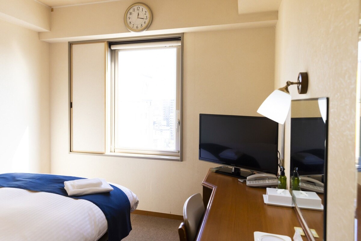 A hotel room with a bed, a desk with a TV, a clock, and a window.