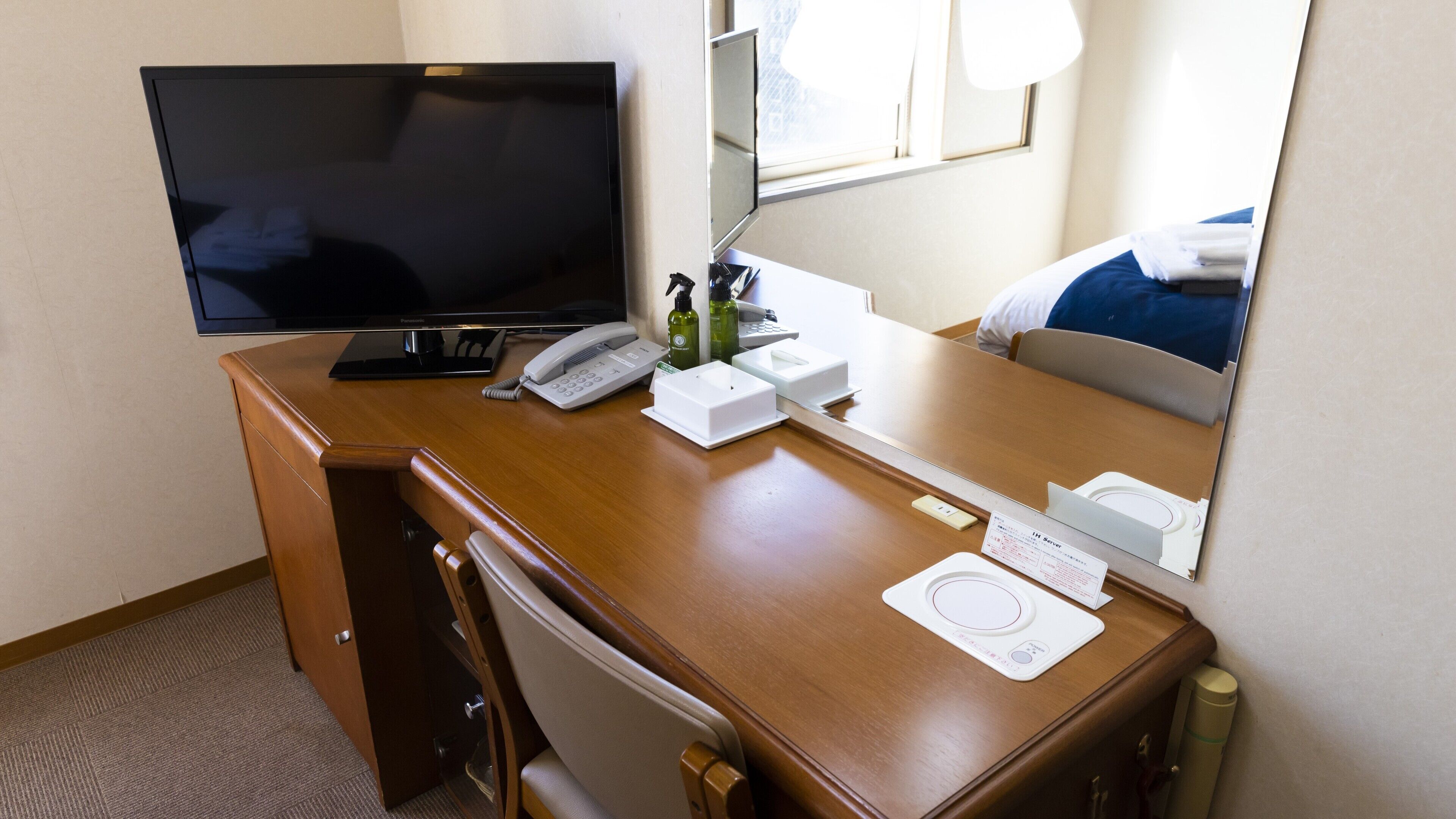 A hotel room with a wooden desk, a flat-screen TV, a telephone, a mirror, a bedside table, and a bed.
