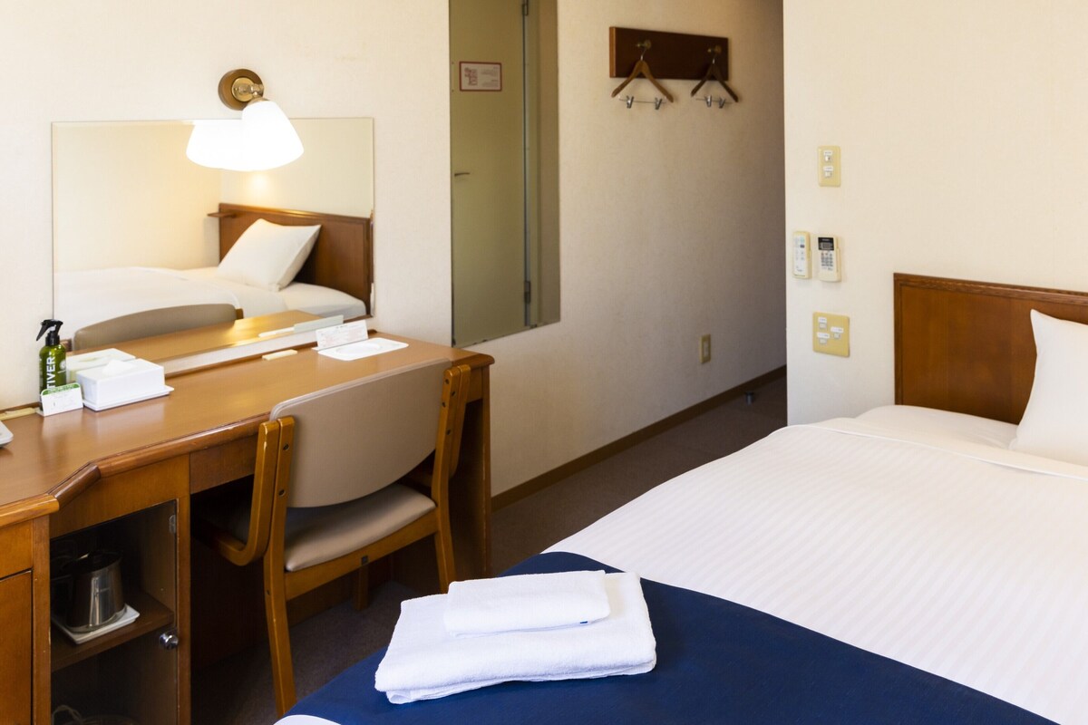 A hotel room with a bed, a desk with a lamp, a chair, a mirror, and a small shelf with toiletries.