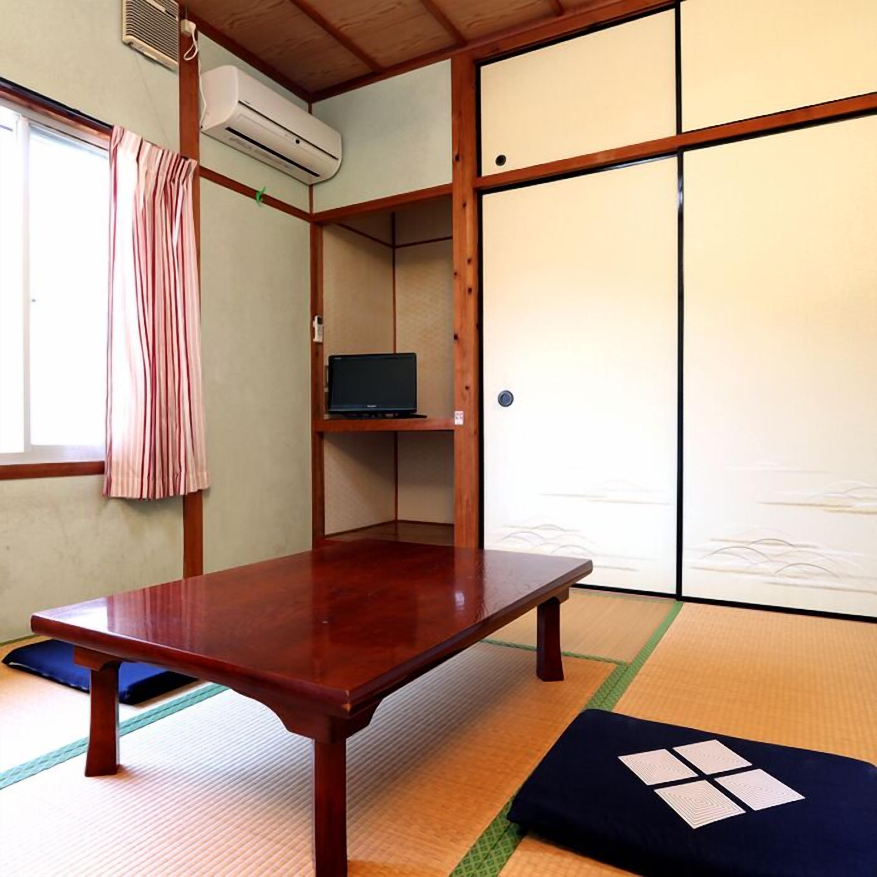 Una habitación japonesa tradicional con suelo de tatami, una mesa de madera, un televisor en un estante y puertas correderas.
