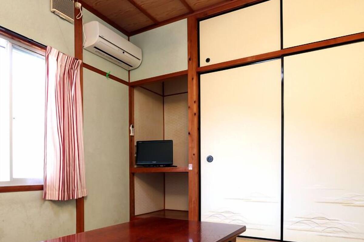 Una habitación japonesa tradicional con suelo de tatami, una mesa de madera, un televisor en un estante y puertas correderas.