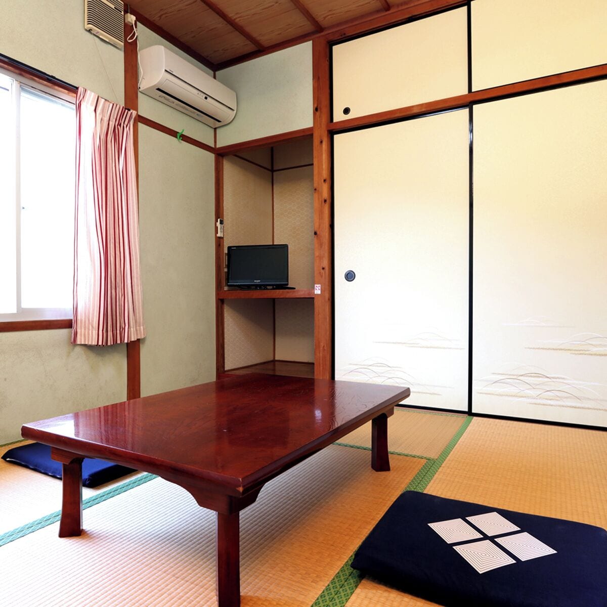 A traditional Japanese room with tatami flooring, a low wooden table, a television on a shelf, and sliding doors.