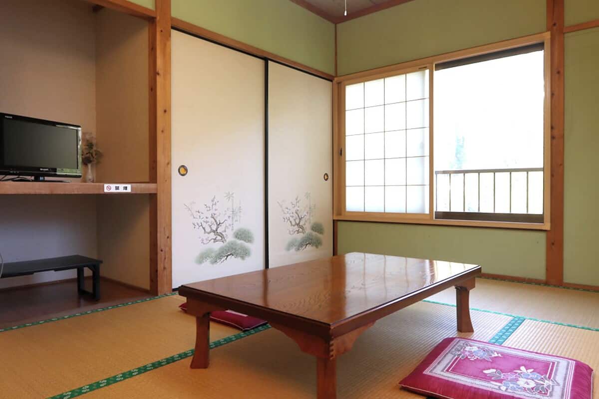A traditional Japanese room with tatami flooring, a low wooden table, sliding doors with floral designs, and a television on a shelf.