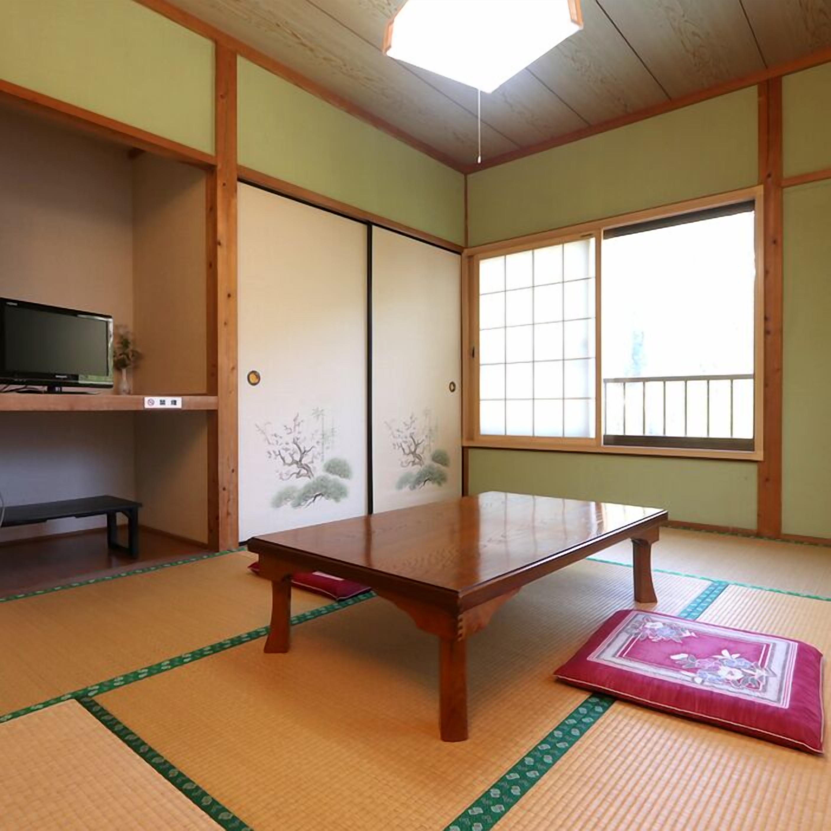Una habitación japonesa tradicional con suelo de tatami, una mesa de madera baja, puertas correderas decoradas y un televisor en un estante, además de ventanas amplias.