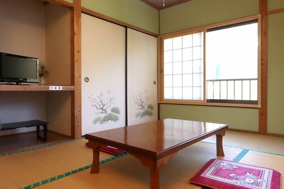 A traditional Japanese room with tatami flooring, a low wooden table, sliding doors with artwork, a television on a shelf, and large windows.