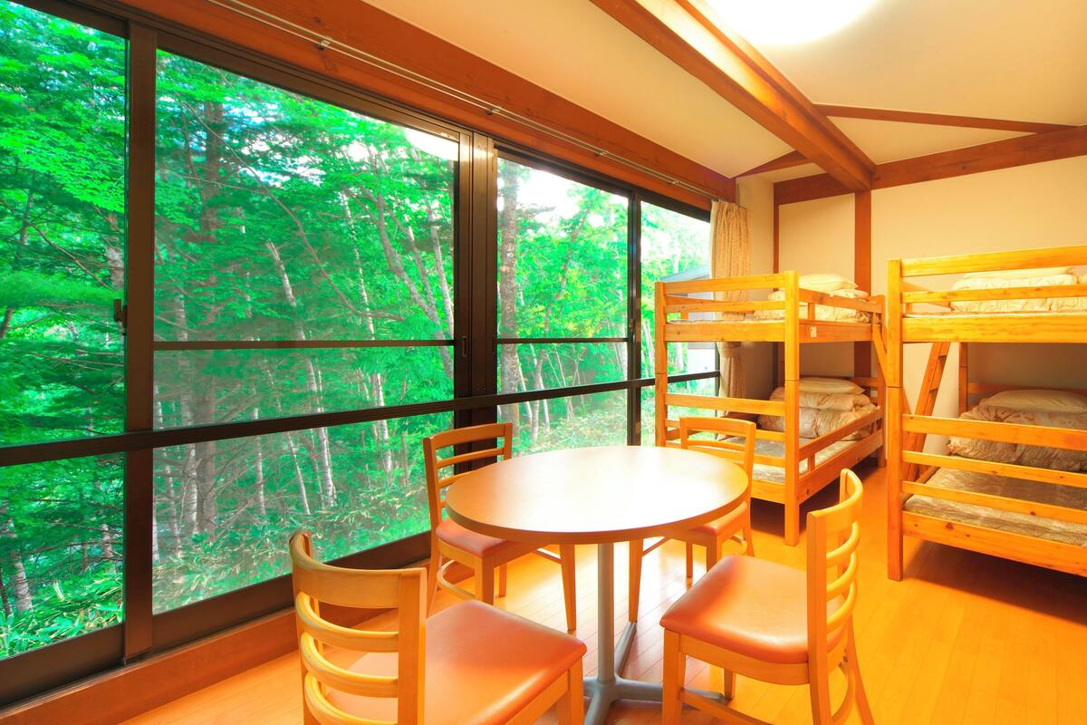 A room with bunk beds, a round table, and chairs, large windows with a forest view.
