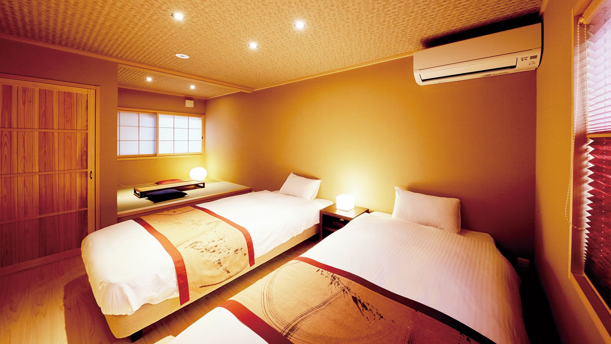 A hotel room with two beds, a wooden wardrobe, a window with blinds, an air conditioning unit, and a bedside table with a lamp.