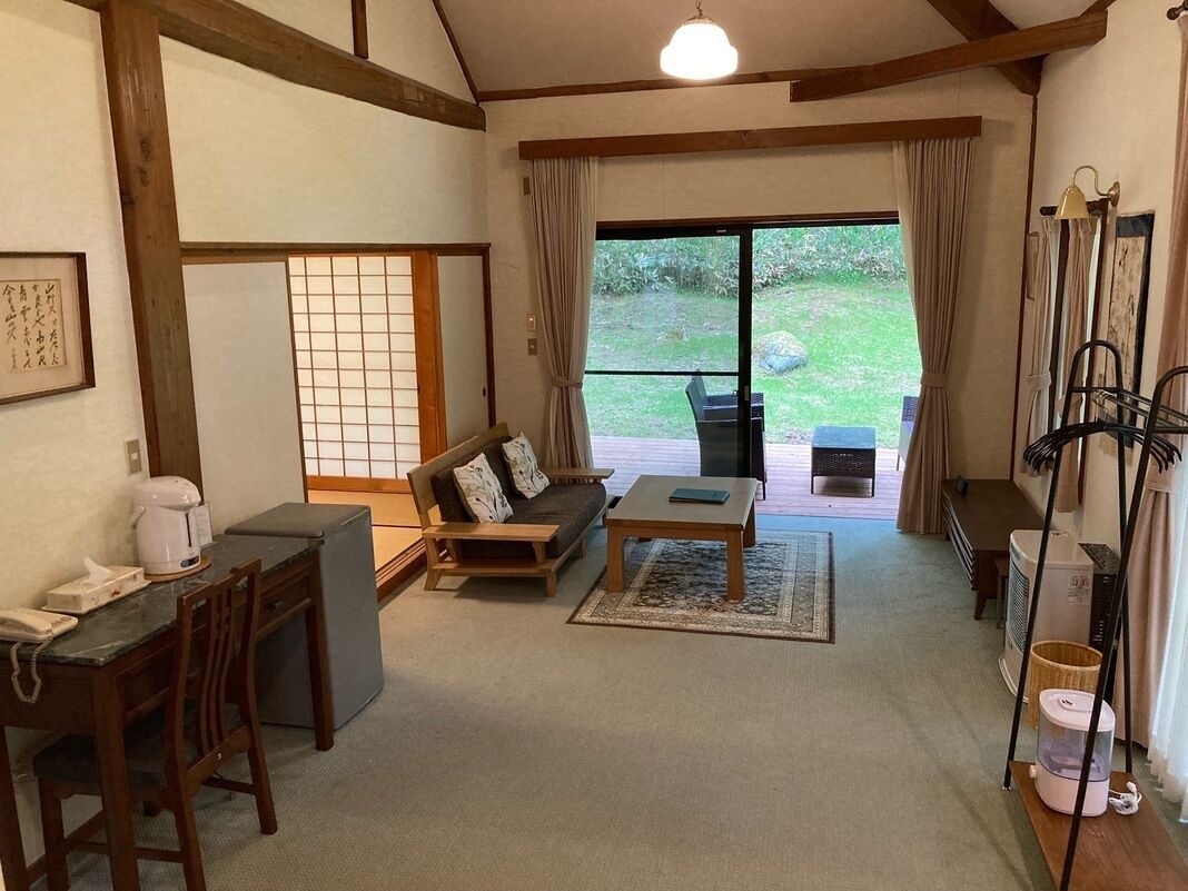 Ein traditioneller japanischer Raum mit Tatami-Boden, einem niedrigen Tisch, einer Couch, einer Schiebeglastür zu einem Garten und einem kleinen Schreibtisch mit Wasserkocher.