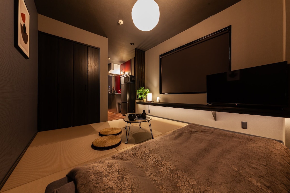 Una habitación compacta con cama, televisión, una mesita con una planta y una zona de cocina al fondo.